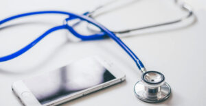 A blue stethoscope and a smartphone lie on a white desk